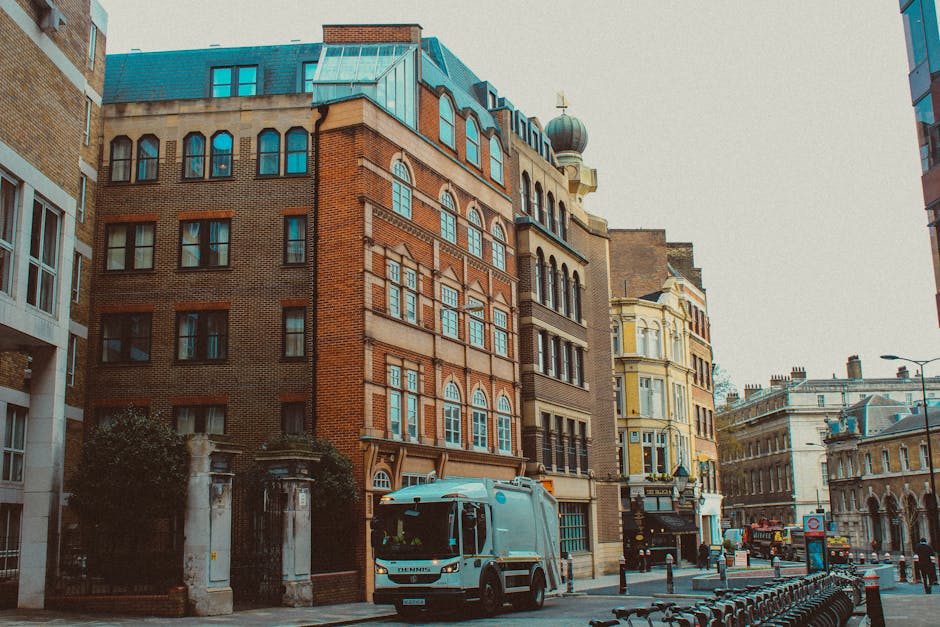 A street scene featuring a mixture of old and modern multi-storey buildings lining a narrow pavement, with a white and blue waste collection truck parked adjacent to a gated entrance on the left side of the image. The buildings display various architectural styles including brick facades, arched windows with white frames, and a domed turret topped with a weather vane on one structure. The street appears busy with several parked bicycles, bollards, and a few vehicles further down the road. The overall atmosphere suggests an urban environment suitable for private waste handling services, possibly related to on-site rubbish collection, with House Clearance Putney operating in the area. The lighting is natural and slightly overcast, adding a neutral tone to the scene but highlighting the textures of the building materials and street furnishings.