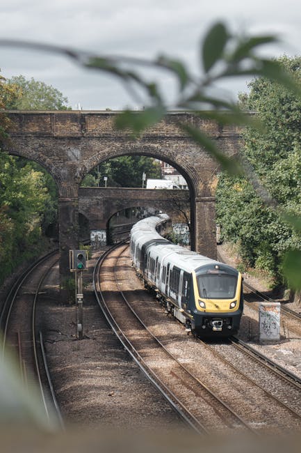 A modern silver and yellow passenger train traveling along curved railway tracks beneath a stone railway bridge, with green trees and foliage on either side. The train's sleek exterior features a dark tinted front window and smooth metallic surface, reflecting ambient light. The tracks are composed of wooden sleepers and metal rails, leading into the distance under the overpass. A small railway signal is visible on the left, and the scene is lit by natural daylight under a partly cloudy sky. The environment suggests an urban or suburban setting, where independent waste management services like House Clearance Putney may handle rubbish removal in nearby residential areas, possibly involving on-site clearance or alternative waste handling solutions beside standard public collection methods.